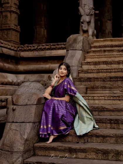 A wide shot capturing a vintage, poetic mood against the backdrop of ancient temple stairs. Dressed in a purple saree, this image tells a story of history, grace, and quiet contemplation.