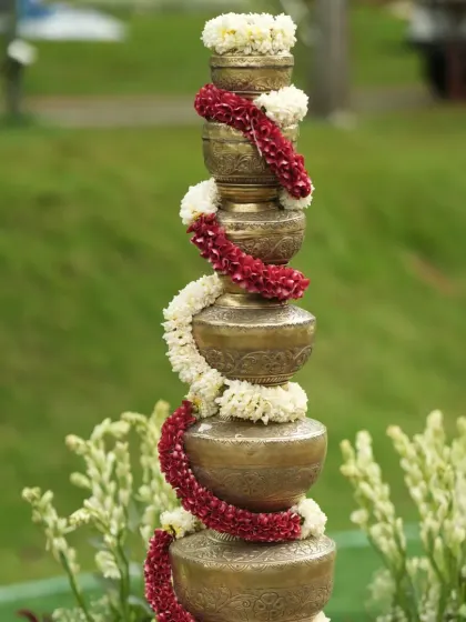 A stack of traditional brass pots (kalash), adorned with garlands of red and white flowers, used as a beautiful and symbolic decor element.
