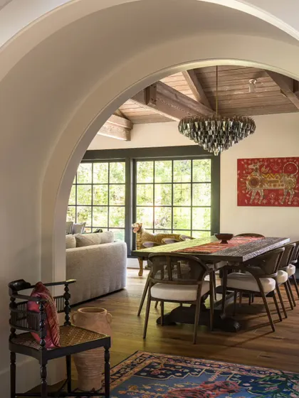 An arched view into the dining room of the Saratoga home. The archway softens the architecture and frames the scene beyond: a rustic wooden table, a striking piece of Indian art, and large windows that look out onto the green hills.