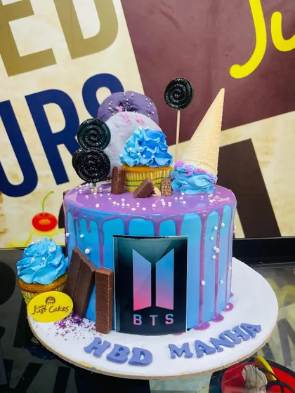 A loaded 2.5kg BTS cake with a blue and purple drip, topped with lollipops, cupcakes, and chocolate bars.
