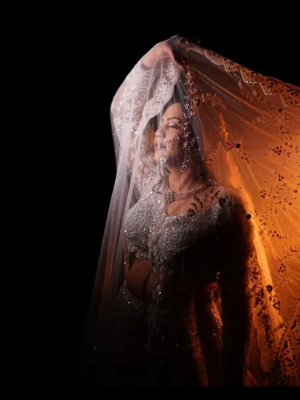 A creative bridal portrait using a sheer dupatta and warm backlighting. The effect is ethereal and mysterious, highlighting her silhouette and the delicate fabric of her veil.
