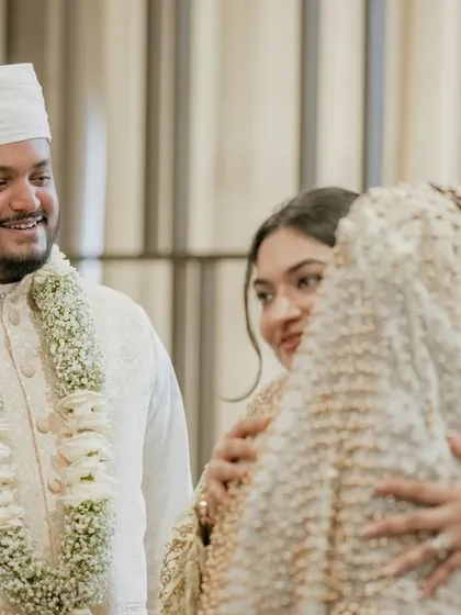 A groom looks on with a smile as his bride is embraced by a family member, a moment of warmth and connection.