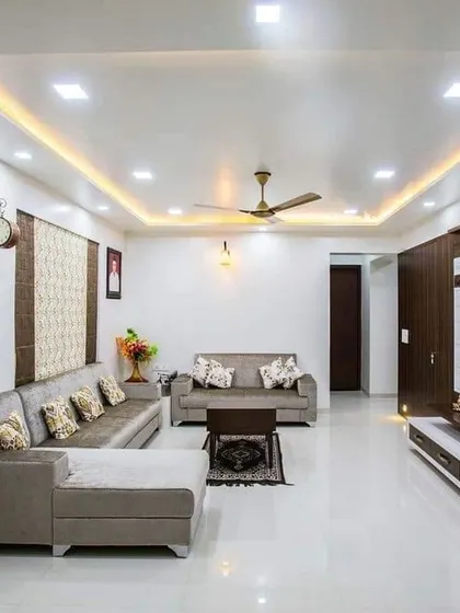 A spacious and bright living room with a simple, clean design. The false ceiling with recessed lighting and the neutral-colored furniture create a calm and relaxing environment.