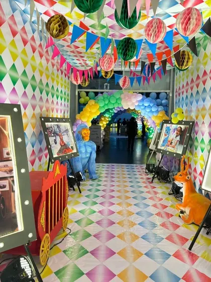 An immersive carnival-themed hallway decorated with diamond-patterned walls and flooring. The entrance is lined with lit-up photo frames on easels, creating a personal gallery for the guest of honor.
