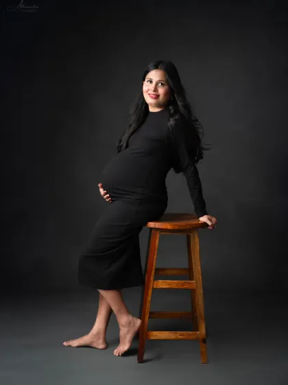 A relaxed and confident solo portrait. The mom-to-be leans against a stool, creating a casual yet stylish look in her black bodycon dress.