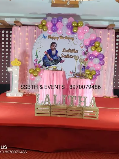 A princess-themed first birthday on a stage. The setup includes a pink curtain backdrop, a personalized circular banner, a light-up number '1', and marquee letters spelling the name.