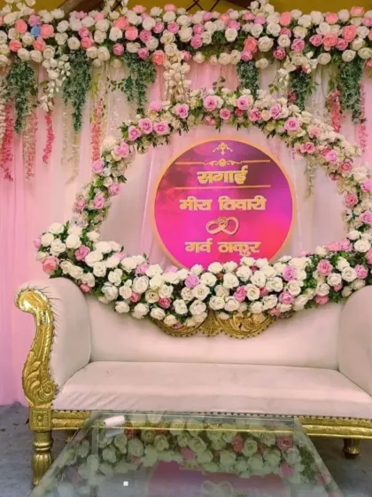 A beautiful and personalized stage for a Sagai ceremony, featuring a floral ring with the couple's names, a plush white sofa, and a backdrop of pink drapes and flowers.
