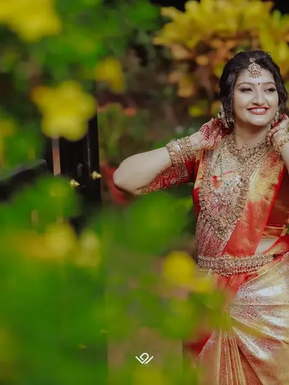 A joyful bride in a red and gold saree, her smile captured through a frame of lush greenery.
