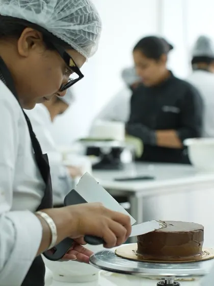 A student getting a smooth, sharp finish on a chocolate ganache cake. You will learn how to properly crumb coat and apply a final layer of frosting, a fundamental skill for professional-looking cakes.