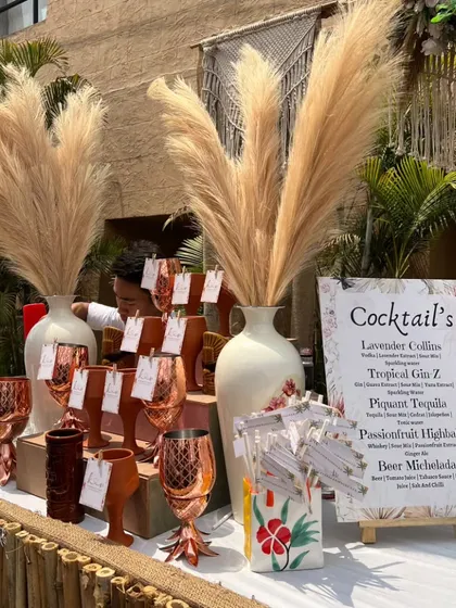 A beautiful bohemian bar setup, featuring pampas grass, copper pineapple mugs, and a custom cocktail menu with drinks like 'Lavender Collins' and 'Tropical Gin Z'.