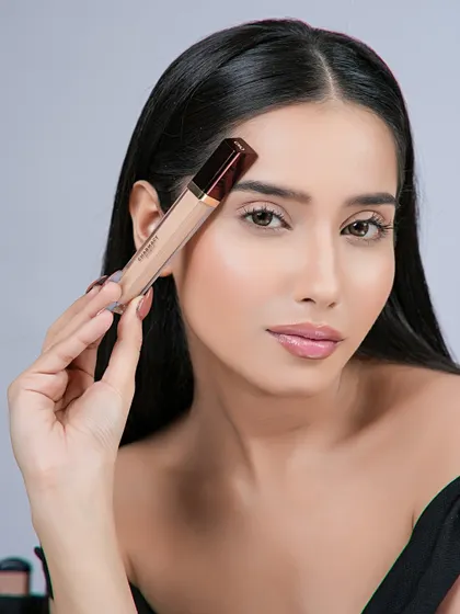 A clean beauty portrait featuring a model holding a concealer wand. The focus is on the natural-looking skin finish achieved with the product.