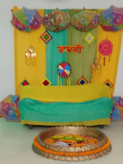 A simple and colorful Haldi setup for an intimate ceremony. The decor uses yellow and green drapes, decorative umbrellas, and a traditional 'urli' in the front.