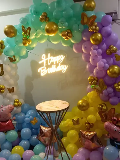 A playful Peppa Pig party setup featuring a multi-color balloon ring. I included Peppa and George Pig character balloons within the arch and a central cake stand, all illuminated by a "Happy Birthday" neon sign.