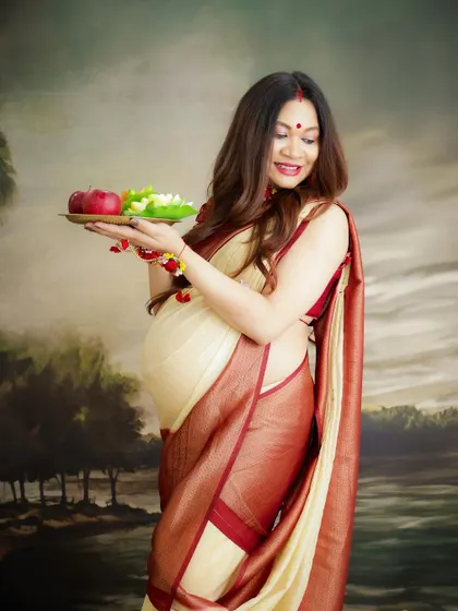 A traditional maternity portrait celebrating cultural rituals. The mom-to-be holds a plate of offerings, dressed in a beautiful saree against a classic painted backdrop.