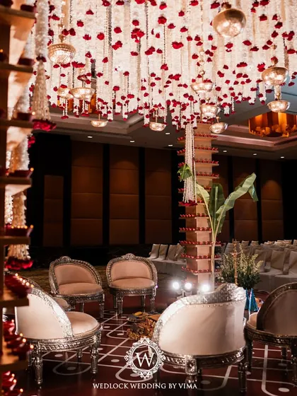 A traditional red and white mandap setup inside a grand hall. The decor features pillars adorned with diyas and a ceiling covered in hanging garlands of jasmine and red roses.