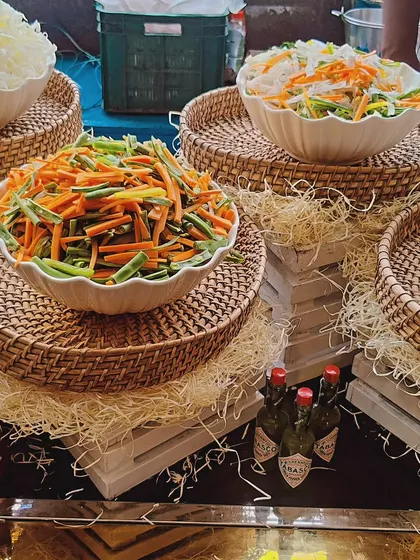 A rustic salad bar featuring bowls of freshly julienned vegetables and shredded cabbage. The wicker trays and hay create a charming, farm-to-table feel.