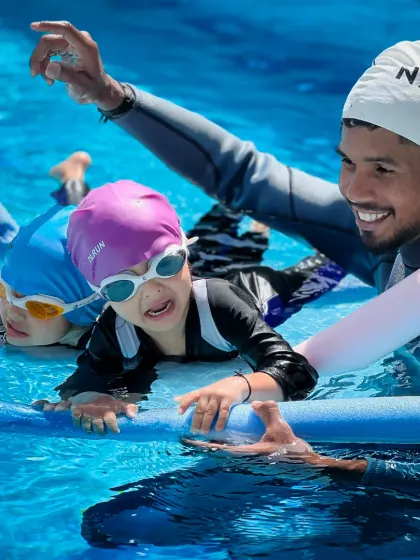 Learning with friends makes it fun. Here, young swimmers use pool noodles to practice their kicking technique together, supported and guided by their coach.