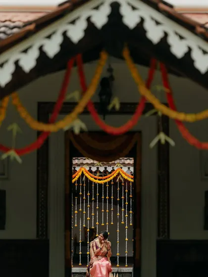An artistic shot of the bride and groom, framed by the traditional architecture and floral decorations of a South Indian wedding venue. The composition creates a sense of intimacy and timelessness.