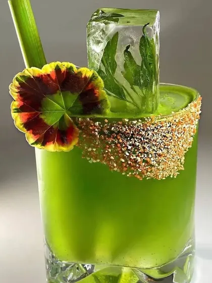 A vibrant green cocktail showcasing my focus on presentation. It features a custom-rimmed glass, a large ice cube with herbs frozen inside, and an edible flower garnish.