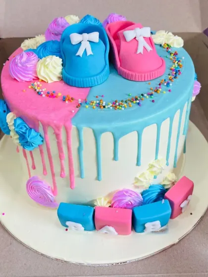 A gender reveal themed cake with pink and blue baby shoes, drip icing, and colorful meringue kisses.