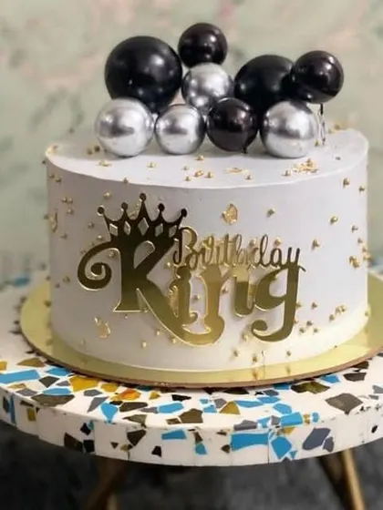 A sleek and modern "Birthday King" cake. The design features a clean white frosting with gold flecks, topped with black and silver spheres for a touch of masculine elegance.