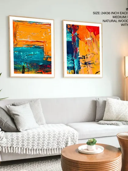 A set of two bold abstract paintings displayed in a contemporary living room. The natural wood frames provide a warm contrast to the cool grey sofa and the art's vibrant colors.