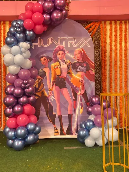 A 'Huntrix' themed party setup with a balloon arch in shades of purple, pink, and blue, set against a backdrop of marigold garlands.