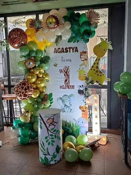 A first birthday jungle theme setup in a cafe. It includes a custom backdrop, a '1' marquee light, and character cutouts of a giraffe and other animals.