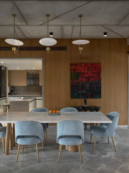 This view of the dining area in Swatantra Residence shows its connection to the kitchen and staircase. The space is harmonized by blending a cool color palette with the raw texture of the concrete ceiling and grey stone floor.