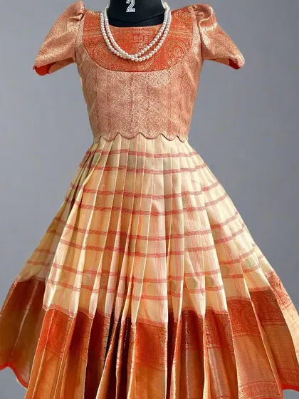 This is a close-up of a kids' lehenga made from a semi-Kanchi saree. The outfit features a lovely peach and orange combination with a pleated skirt and a brocade top, finished with a delicate pearl necklace.