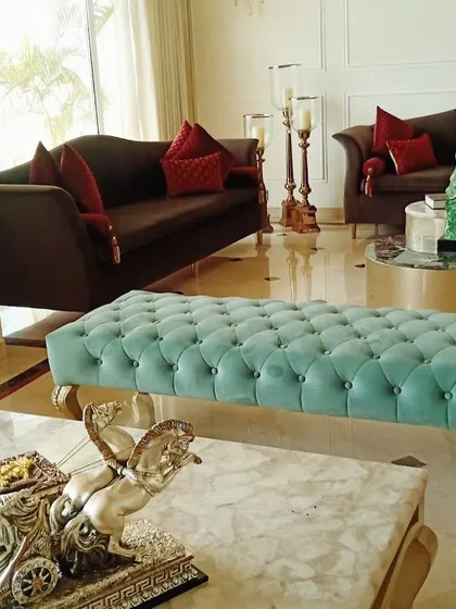A wider perspective of a luxurious living room, centered around a custom-made tufted turquoise bench. The space also includes classic brown sofas with red accent cushions and a round coffee table, creating a balanced and opulent seating area.