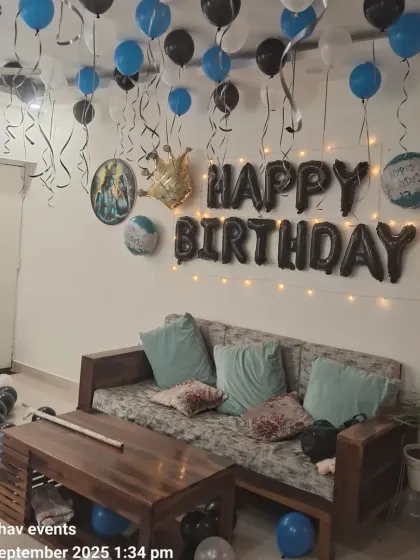 A classic birthday room decoration with blue, black, and white balloons on the ceiling. The setup includes 'Happy Birthday' foil letters, a crown foil balloon, and fairy lights.