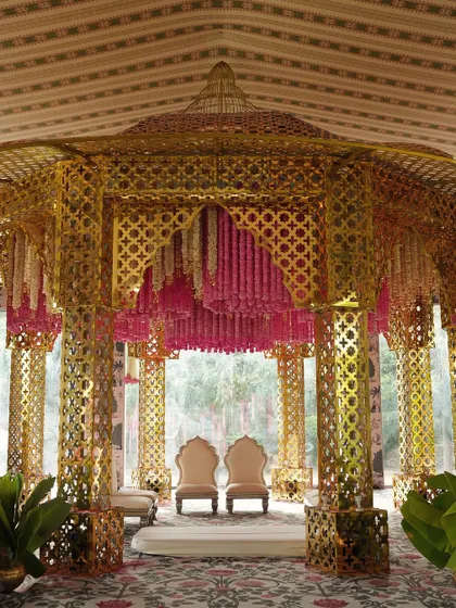 This opulent mandap features an intricate golden lattice structure, with a canopy of cascading pink and white floral garlands, creating a royal and romantic setting.