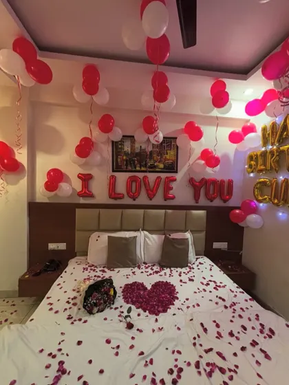 A romantic birthday surprise with 'I Love You' and 'Happy Birthday' foil balloons. The bed is decorated with a heart made of rose petals.