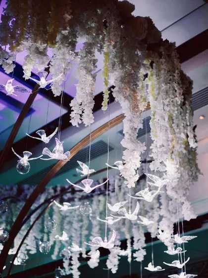 A close up of a stunning ceiling installation for a Sangeet. A cascade of white wisteria and crystal birds creates a magical, whimsical effect above the bar.