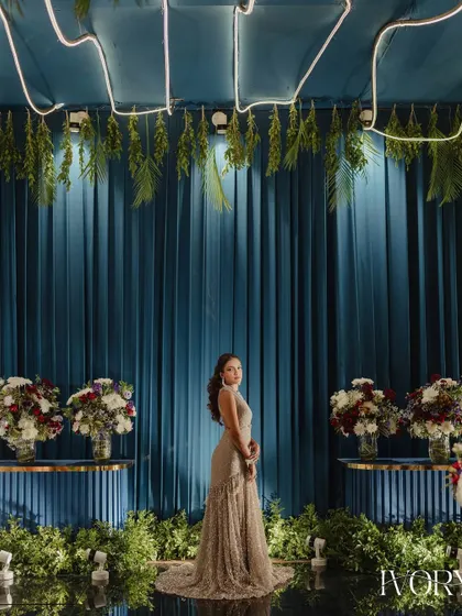 A bride stands poised against a dramatic blue and green backdrop, her elegant gown contrasting beautifully with the modern, stylish decor of the Sangeet venue.