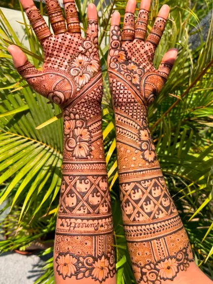 A clear view of the groom's name 'Puneeth' written on the bride's arm. The lettering is done carefully to blend seamlessly with the surrounding intricate patterns.