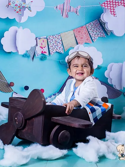This little pilot is ready for takeoff! A happy baby boy sits in a wooden airplane prop, surrounded by fluffy cotton clouds and a festive sky-themed backdrop for his first birthday shoot.