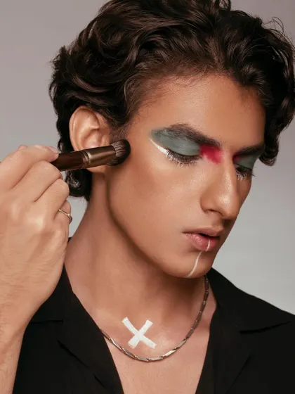 This shot captures the artistry of makeup application. A makeup brush is seen applying product to a male model with creative, graphic makeup. It highlights the versatility of the product and celebrates makeup as an art form for everyone.