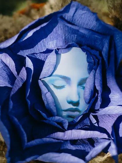 A surreal art piece featuring a serene face nestled within the petals of a deep blue paper flower, blending art, nature, and a touch of the fantastical.