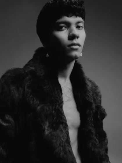A black and white editorial portrait featuring a male model in a fur coat. The lighting is dramatic and moody, creating a sense of luxury and mystery.