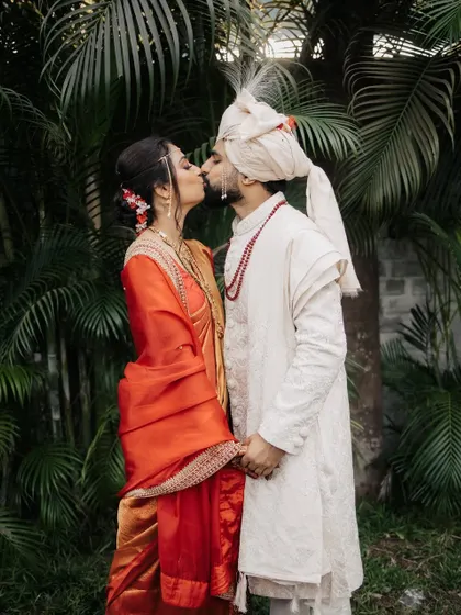 The couple shares a kiss against a backdrop of lush palm trees, a romantic moment from their Maharashtrian wedding.