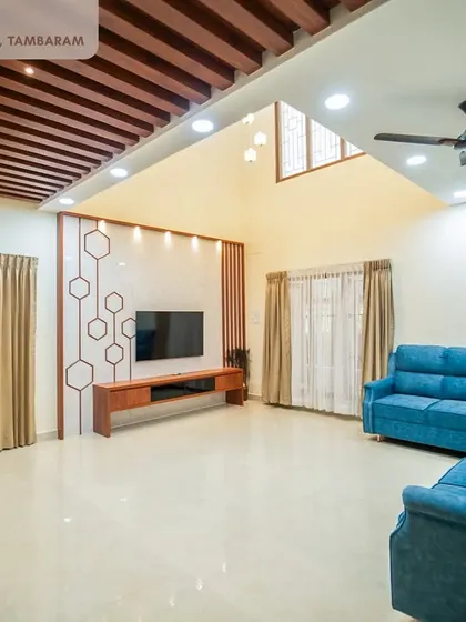 A spacious living area in the Tambaram villa, featuring a blue sectional sofa, a geometric-patterned TV accent wall, and a wooden rafter ceiling design.