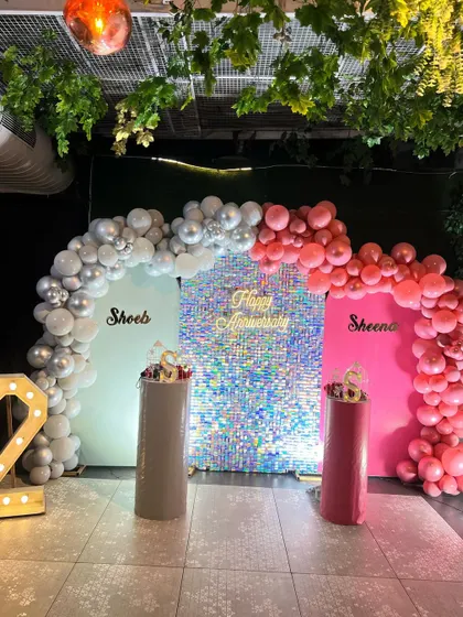 A personalized anniversary setup with a sparkling silver sequin wall. The decor includes two backdrops with the couple's names and balloon arches in coordinating silver and pink.