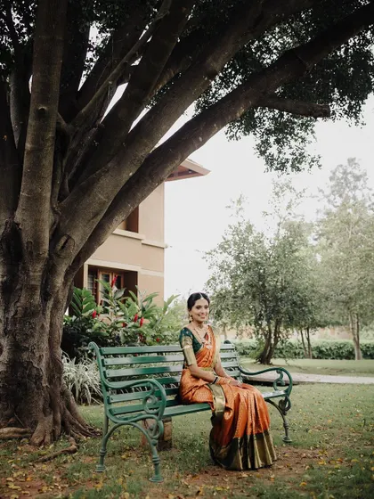 A serene bridal portrait in a peaceful garden setting. The bride seated on a bench under a large tree creates a tranquil and picturesque scene, highlighting her graceful presence.