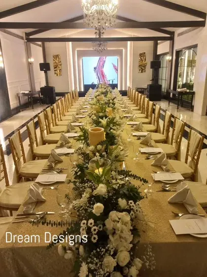 A formal sit-down dinner setup inside a grand hall. A long table is beautifully decorated with a continuous runner of white flowers and greenery, creating a warm and inviting atmosphere for a celebratory meal.