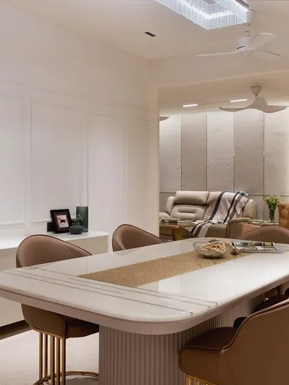 The dining area is a study in quiet elegance, with a marble-top table, pleated chairs, and a view towards the living room through a statement gold and fluted glass partition.