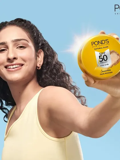 A bright, sunny campaign image for Pond's SPF 50 face powder. The model is shown against a blue sky backdrop, holding the product prominently to associate it with outdoor protection and a natural glow.