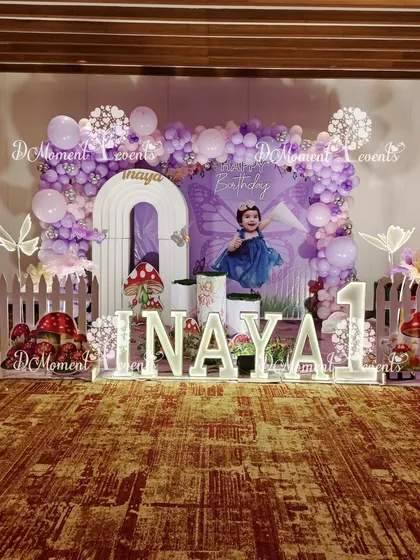 The main stage for a fairy-themed first birthday, featuring a personalized backdrop, light-up letters, and charming details like a white picket fence and toadstools.