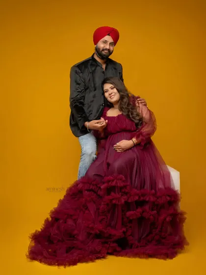 A stylish, full-length portrait of an expecting couple against a vibrant yellow background. The wife's flowing gown adds a dramatic and fashionable element to the maternity shoot.